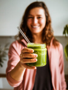 Frau unscharf im Hintergrund hält grünen Smoothie in die Kamera